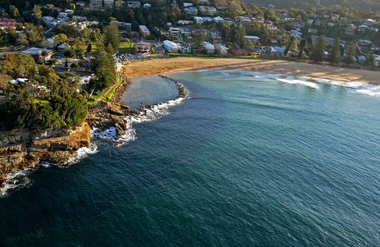 Avoca Beach from above, the lagoon mouth, headland, and houses under the trees.