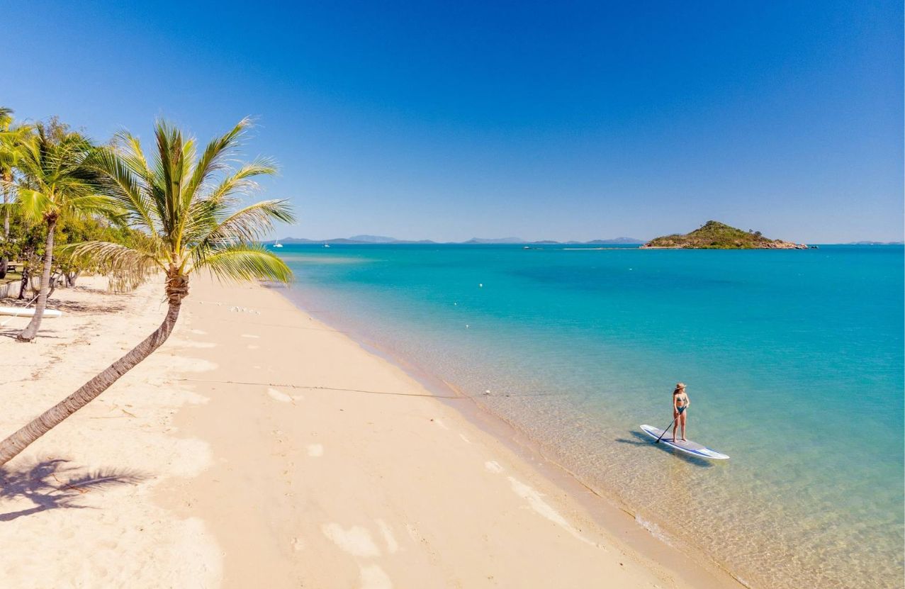 Hydeaway Bay, a woman on a paddleboard in knee-deep turquoise water beside leaning palms.