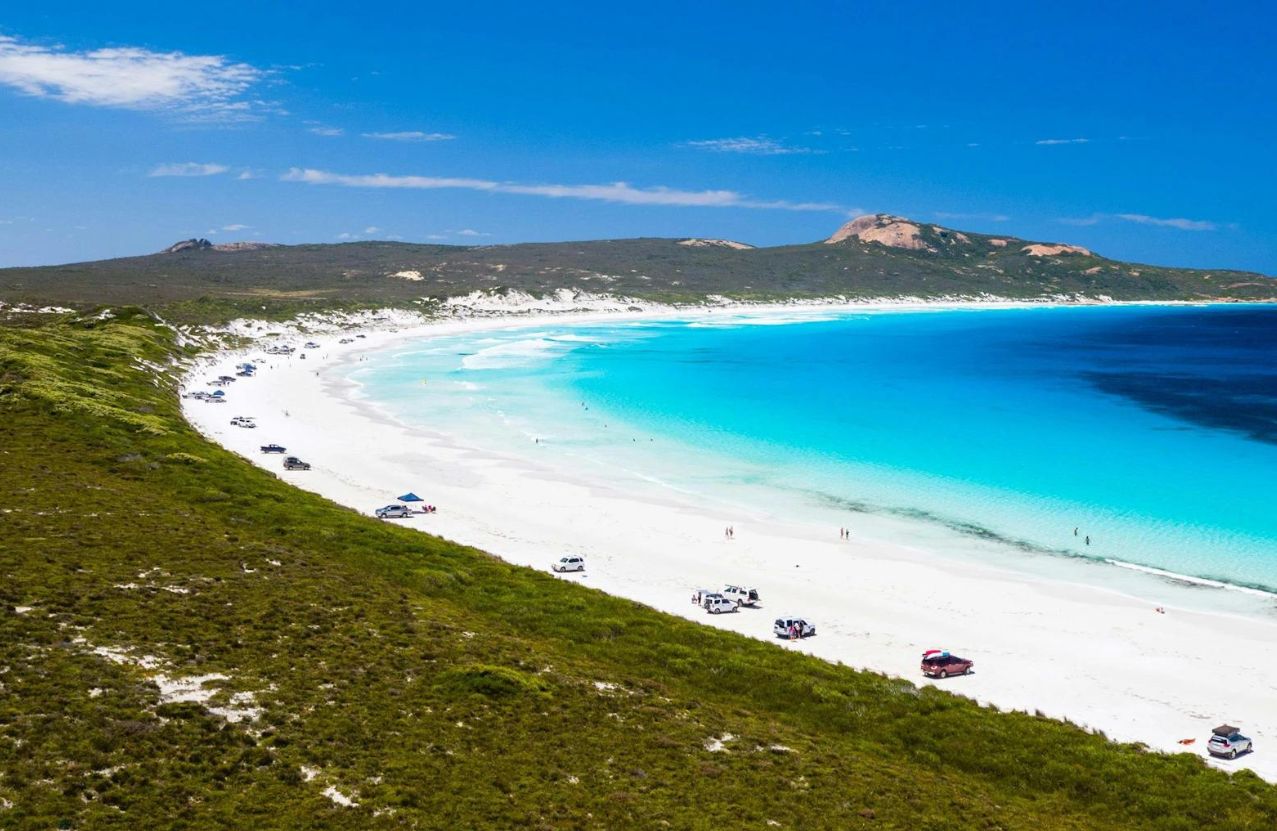 Lucky Bay, cars parked on white sand beside electric-blue water and granite peaks.