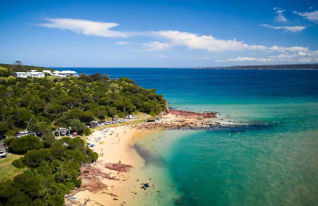 Merimbula Bar Beach, a sandy point, swimmers, and deep blue water past the rocks.