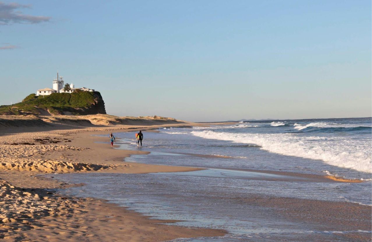 Two surfers walking the wet sand at Nobbys Beach, the lighthouse on the headland.
