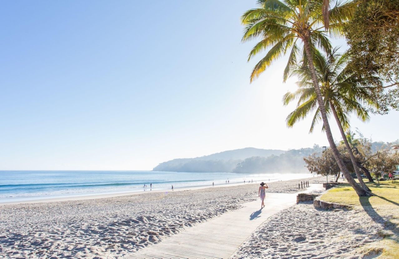 Noosa Main Beach, a boardwalk, leaning palms, and a walker in the morning glare.
